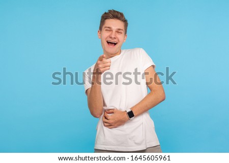 Haha, look at you! Portrait of amused man in casual white t-shirt holding belly, bursting into laughing and pointing to camera, mocking taunting people. indoor studio shot isolated on blue background
