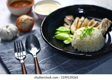 Hainanese Chicken Rice Served On A Black Plate With Dipping Sauce And Hot Broth On White Table.