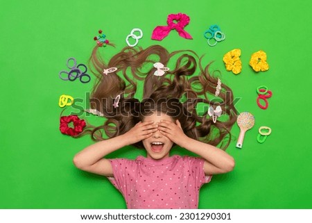 Hair accessories. A happy little girl lies surrounded by elastic bands and hair clips, and covers her eyes with her hands, smiling broadly. Hairstyles for children. Green isolated background.