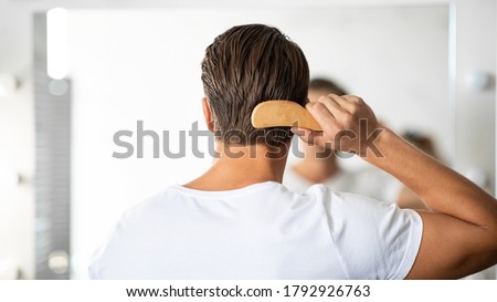 Hair Care. Close up rear view of young man hairbrushing with comb, looking in mirror