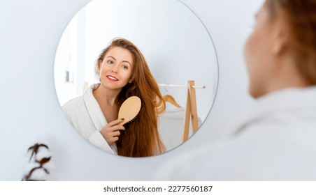 Hair care concept. Happy attractive young redhead woman brushing her thick beautiful hair with comb standing near mirror in bathroom. - Powered by Shutterstock