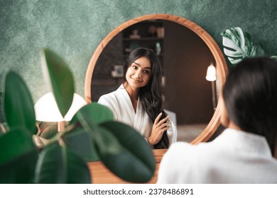 Hair care and self-care with beautiful Indian woman looking in mirror touching her healthy long hair sitting at the dressing table. - Powered by Shutterstock