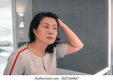Hair loss mature Asian woman touching her hair styling or coloring gray hair looking at herself in home mirror. Beautifiul chinese professional lady makeup.