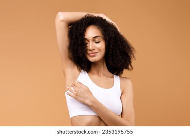 Hair removal concept. Happy latin lady demonstrating her armpit with smooth skin and smiling, posing in white underwear over brown background, copy space