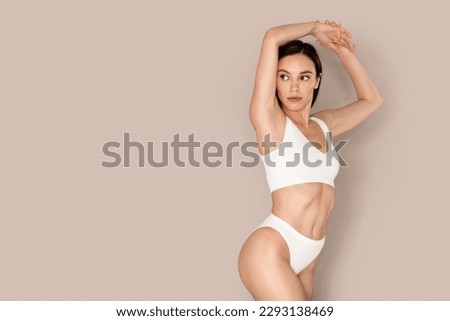 Hair removing, epilation. Attractive brunette young woman in white comfy underwear raising hand up, showing clean hairless armpit, looking at copy space for advertisement, beige studio background