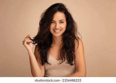 Coiffure, concept de coiffure. Superbe femme millénaire orientale en haut beige posant sur fond studio, montrant ses beaux cheveux longs après le traitement des cheveux du salon et souriant à la caméra : photo de stock