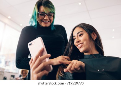 Hair stylist in colorful hairstyle looking at mobile phone while working on woman's hair. Woman showing her preferences to the hairstylist in mobile phone at the salon.