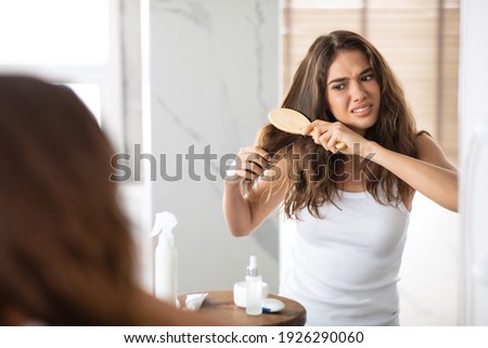 Hair Tangling Problem. Unhappy Young Woman Untangling And Brushing Her Long Tangled Hair Standing Near Mirror In Bathroom At Home. Damaged Hair Combing And Beauty Treatment Concept. Selective Focus