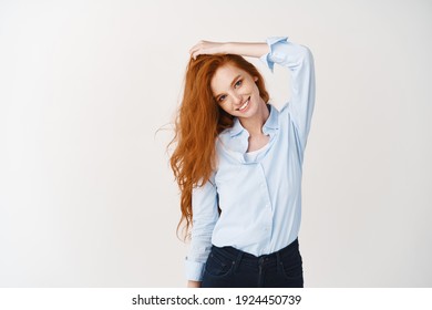 Haircare and beauty concept. Young woman with blue eyes and freckles touching shiny, healthy red hair and smiling at camera, white background.