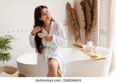 Haircare. Brunette Female Brushing Hair With Hairbrush Detangling Split Ends Smiling Sitting On Bathtub Wearing Bathrobe In Modern Bathroom At Home. Beauty Routine Concept