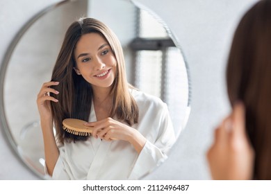Haircare Concept. Attractive Young Lady Brushing Her Thick Beautiful Hair With Comb While Standing Near Mirror In Bathroom, Happy Woman Wearing White Silk Robe Looking To Her Reflection And Smiling - Shutterstock ID 2112496487