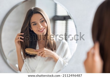 Haircare Concept. Attractive Young Lady Brushing Her Thick Beautiful Hair With Comb While Standing Near Mirror In Bathroom, Happy Woman Wearing White Silk Robe Looking To Her Reflection And Smiling Сток-фото © 