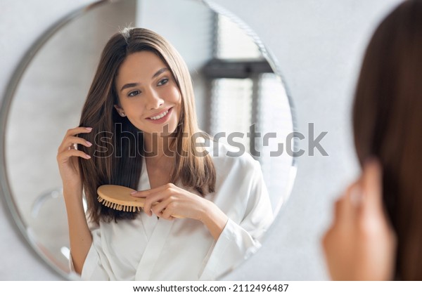 Haircare Concept. Attractive Young Lady Brushing\
Her Thick Beautiful Hair With Comb While Standing Near Mirror In\
Bathroom, Happy Woman Wearing White Silk Robe Looking To Her\
Reflection And Smiling