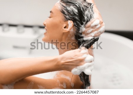 Haircare Cosmetics. Woman Washing Head Applying Shampoo On Long Brown Hair Posing With Eyes Closed Sitting In Bathtub In Modern Bathroom At Home. Side View, Selective Focus