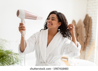 Haircare. Happy Black Woman Drying Curly Hair With Hairdryer Standing In Modern Bathroom Indoors, Wearing Bathrobe. Female Enjoying Hair Styling Routine Concept