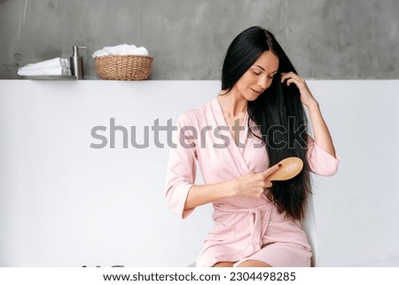 Haircare. Pretty charming caucasian brunette long haired woman, in a pastel pink dressing gown, sitting in a bathroom at home, brushing her thick black hair with wood comb, looking at hair, smiling