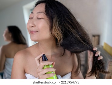 Haircare. Smiling Asian Woman Applying Hair Spray To Split Ends, Young Happy Female Standing Wrapped In Towel After Shower Using Avocado Conditioner At Home.