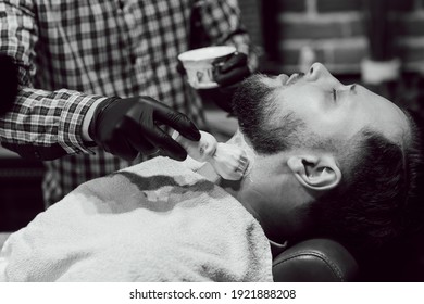 Haircut In The Barbershop. Black And White Close Up View Of Young Bearded Man Getting Haircut While Sitting In Chair At Barbershop. Hairdresser Lubricates The Beard With Shaving Gel