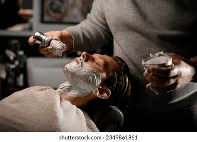 Hairdresser applying shaving foam on client's beard before shaving in barber shop. Side view. Man's service - Powered by Shutterstock