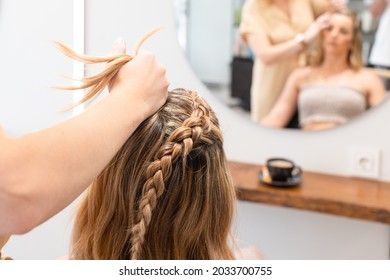 Hairdresser Braiding A Woman In Her Blonde Hair In A Hair Salon. View From Behind. Selective Focus