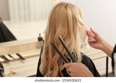 Hairdresser curling woman's hair with flat iron in salon, closeup
