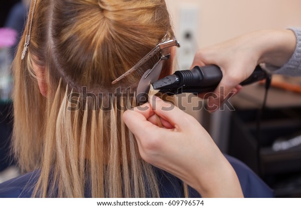 The hairdresser\
does hair extensions to a young girl, a blonde in a beauty salon.\
Professional hair care.