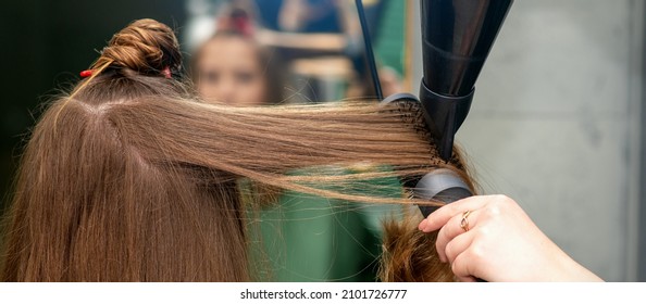 A hairdresser is drying long brown hair with a hairdryer and round brush in a hairdressing salon.