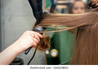 A hairdresser is drying long brown hair with a hairdryer and round brush in a hairdressing salon.