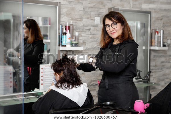 Hairdresser dyeing the hair\
of a brunette woman.  Workplace with dividing plastic screens for\
safety distance between clients. New normal during coronavirus\
pandemic