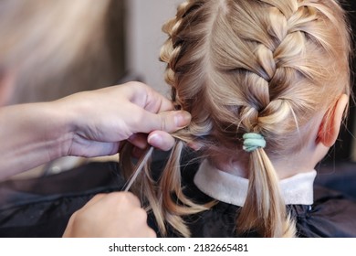 Hairdresser makes hairdo braids for young baby in barber shop. Barber woman make fashionable hairstyle for cute little blond girl child in modern barbershop, hair salon. Concept hairstyle and beauty