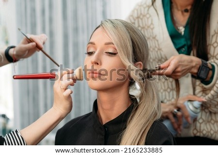 A hairdresser and a makeup artist together prepare a beautiful model for a photo shoot. The girl gets her hair and makeup done in a beauty salon.