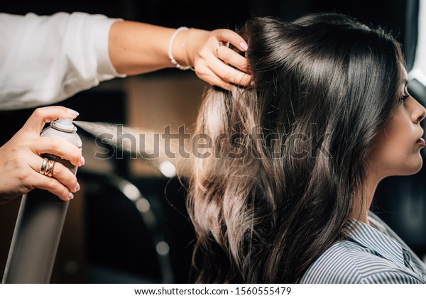 Hairdresser spraying woman’s long black hair with\
hair spray.