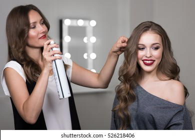 Hairdresser Using Hairspray On Client's Hair At Salon, Portrait Of Two Beautiful Women
