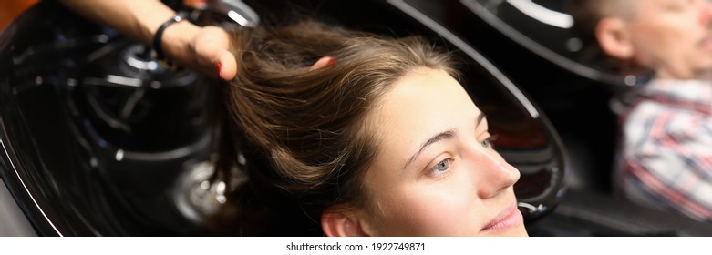 Hairdresser wash hair to client in barber shop. Beautiful woman sit on chair in cape and with head thrown back and smile. Handsome man wait for wash hair in background. Professional hair and scalp