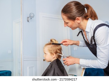 Hairdresser Woman With Comb And Scissors Working On Haircut Of A Little Blond Boy Profile View