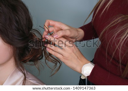 Hairdresser at work in process of creating a wedding hairstyle. Master fixes lower topknot of hair with invisible hairpins and hat pins. Hand hairdresser straightens brown curls. Beauty industry