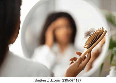 Hairloss Concept. Upset Black Woman Holding Comb Full Of Fallen Hair After Brushing, Closeup Shot Of African American Female With Brush In Hand Standing Near Mirror, Suffering Alopecia Problem