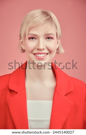 Hairs and makeup. An attractive young girl with a stylish blonde short haircut and bright pink makeup poses in a red jacket on a pink studio background. Makeup cosmetics and hair styling products.