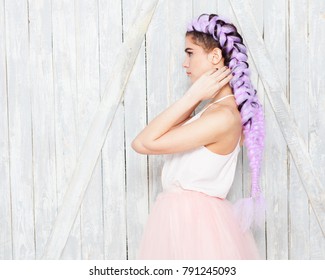 Hairstyle with colored, purple braids. Portrait young cute girl standing in profile showing braids, posing on light wooden background. Summer light outfit. Indoor.