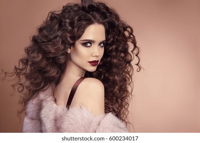 Hairstyle. Fashion brunette girl with Long curly hair, beauty makeup. Glamour portrait of beautiful brunette with marsala matte lips in pink fur coat isolated on beige background.