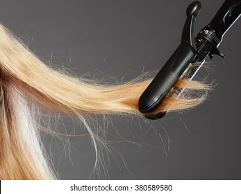 Hairstyling. Closeup Blonde Woman Long Haired Making Hairstyle Hairdo With Electric Hair Iron Straightener Gray Background