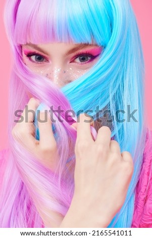 Hairstyling, hair dye. Portrait of a beautiful girl with bright pink makeup hiding her face with her colored purple-blue hair and smiles. Pink background. Beauty concept.