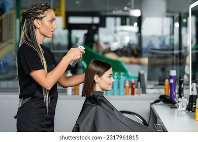 hairstylist spraying hair of female client, hairdresser with braids holding spray bottle near woman with short brunette hair in salon, hair cut, hair make over, side view - Powered by Shutterstock