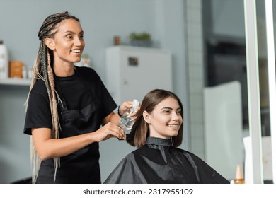 hairstylist spraying hair of happy woman, hairdresser with braids holding spray bottle near female client with short brunette hair in salon, hair treatment, hair make over, hairdo - Powered by Shutterstock