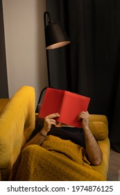 Hairy Arms Guy Reads Book Liyng On Comfortable Yellow Sofa. No Face Male Reader Reading Detective Or Historic Novel At Home Comfy Couch Covered With Plaid Blanket In Stylish Minimalist Interior