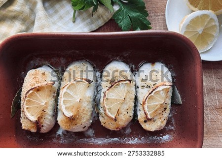 Hake or cod fillet hearts, cooked in a pan in the oven, garnished with lemon slices and aromatic herbs. Seafood. Overhead view.