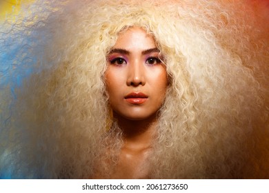 Half Body Portrait Of 20s Asian Caucasian Beautiful Woman Blonde Afro Hair High Fashion Make Up. Cute Girl Smile Look At Camera Over Pastel Red Blue Yellow Colorful Sky Cloud In Studio