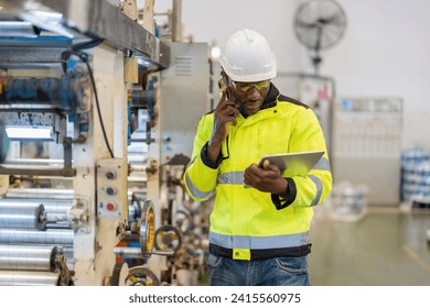 Half body portrait. African male engineer. Using a smartphone to talk to work, holding a laptop, wearing a vest and helmet, next to a working machine at a large company's plastic and steel industry. - Powered by Shutterstock