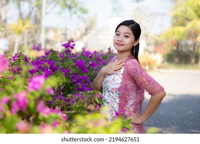 Half Body Portrait Of An Asian Woman Smiling In White Blouse And Pink Blazer In A Park With Purple And Pink Flowers Background.