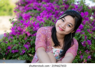 Half Body Portrait Of An Asian Woman Smiling In White Blouse And Pink Blazer In A Park With Purple And Pink Flowers Background.